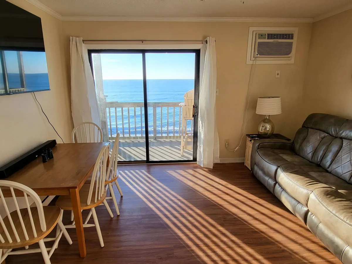 This image captures the open-concept living area with a cozy sofa and a dining table, all oriented toward broad sliding glass doors that frame a sweeping ocean view. Natural light floods the space, and you can see the private balcony just beyond the doors, reinforcing the direct connection between the living space and the sea.