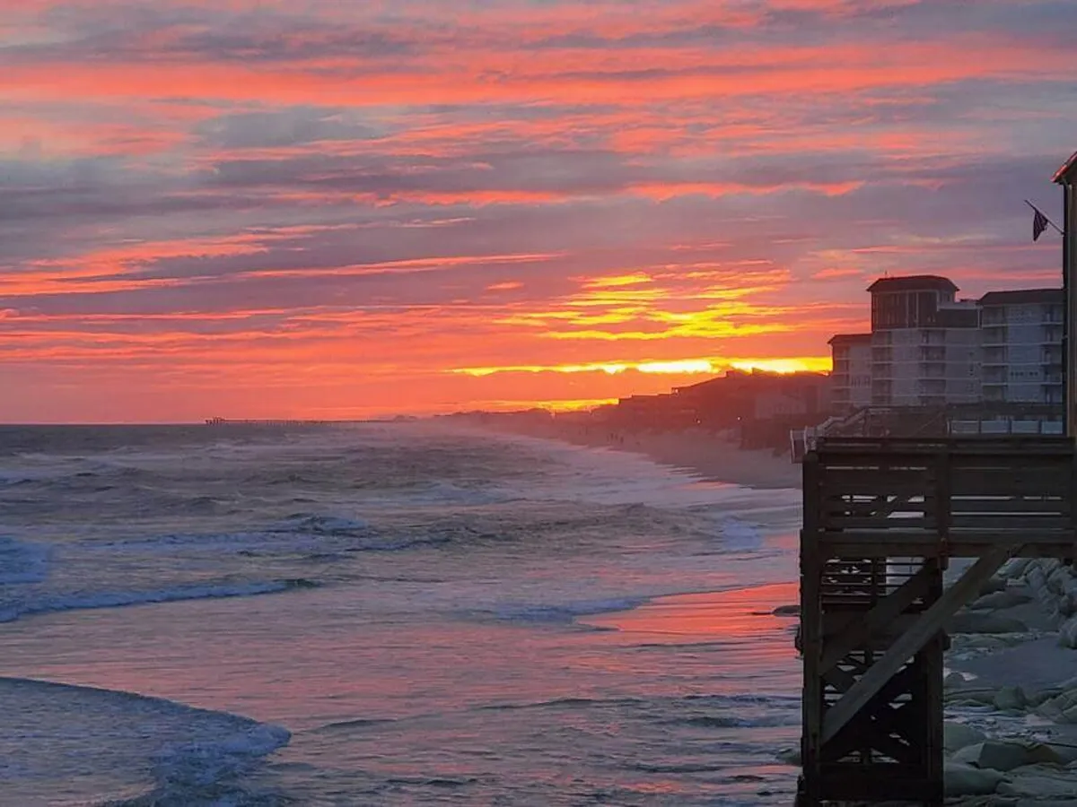 A vibrant sunset casts colorful light over the ocean, with a pier structure extending out and silhouetted buildings framing the horizon. The scene suggests opportunities for fishing, strolling, or serene contemplation at day's end.