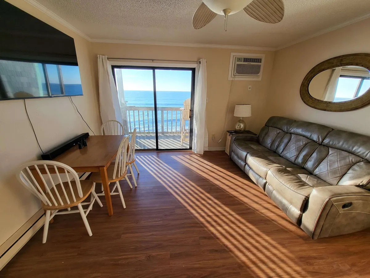 The scene shows a comfortable leather sofa positioned by a large window with a direct view of the ocean, as well as a dining table that overlooks the water. The setup exudes coziness and tranquility, inviting guests to settle in and savor the scenery from inside.
