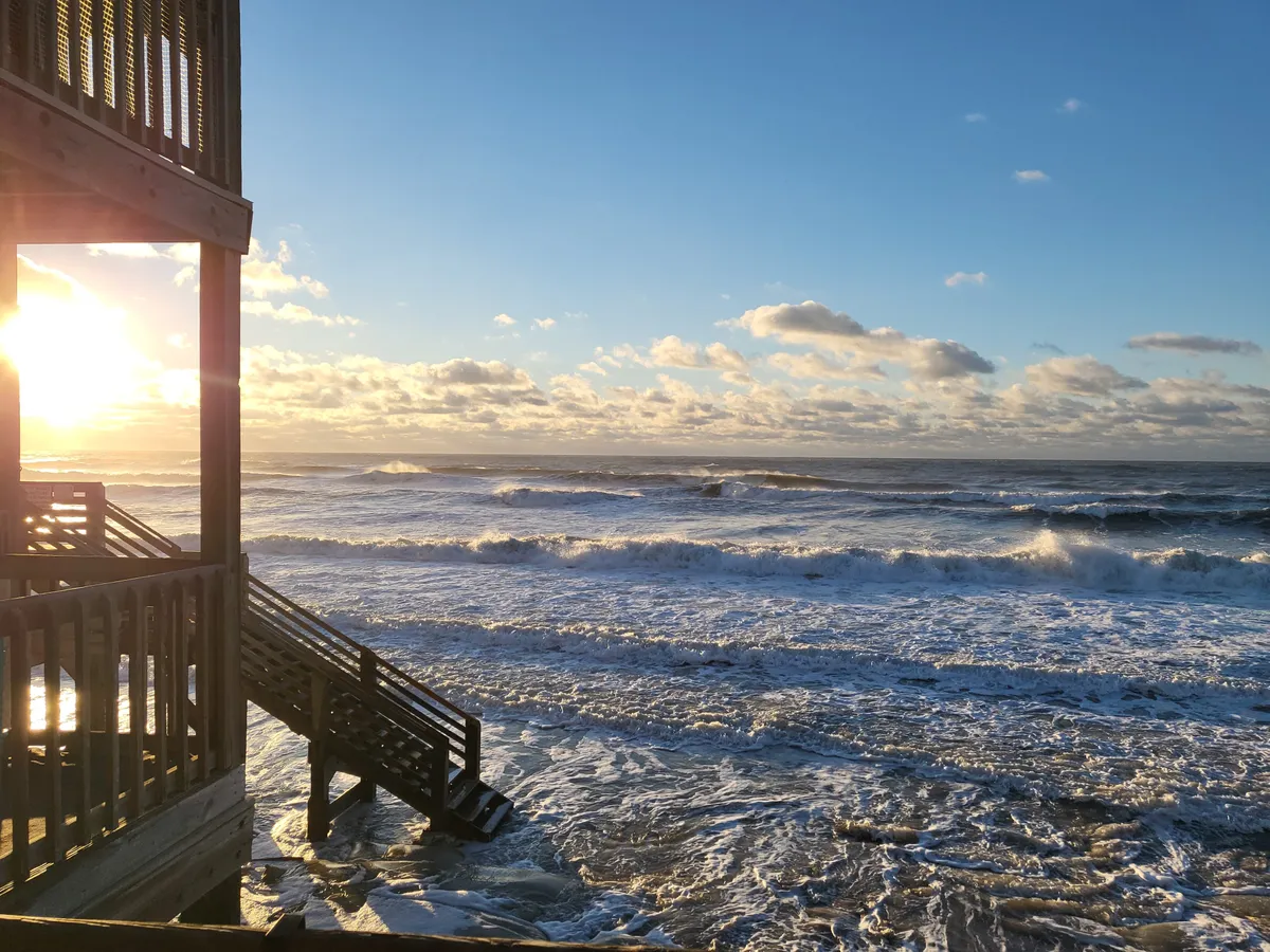 A peaceful sunset paints the sky over the ocean, with gentle waves crashing on the shore and stairs leading down to the beach. The scene captures a serene, romantic ambiance, with natural beauty inviting sunset strolls and reflective moments.