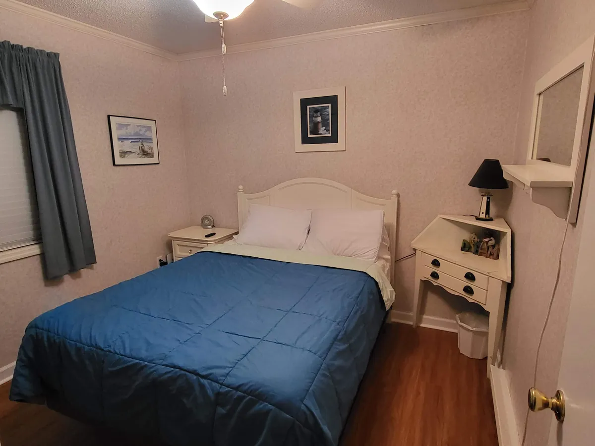 This image shows a serene bedroom with a bed featuring a blue quilt, a nightstand with a lamp, and framed pictures on the wall. Soft natural light fills the room, creating a calm and inviting atmosphere perfect for relaxation.