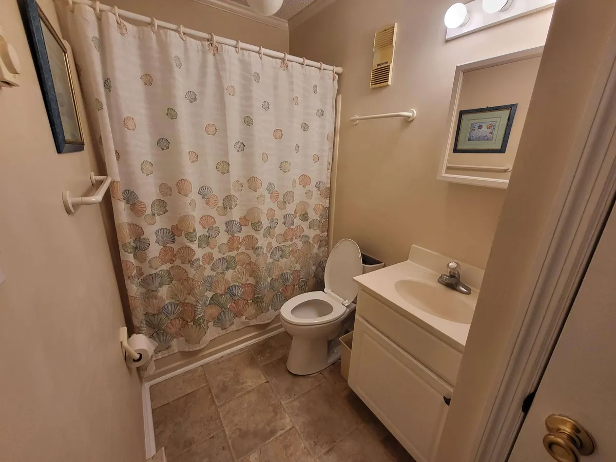 A bathroom interior featuring a shell-patterned shower curtain, a sink with a mirror, and a toilet. The space is bright and clean, with subtle coastal decor touches that create a refreshing and modern vibe.