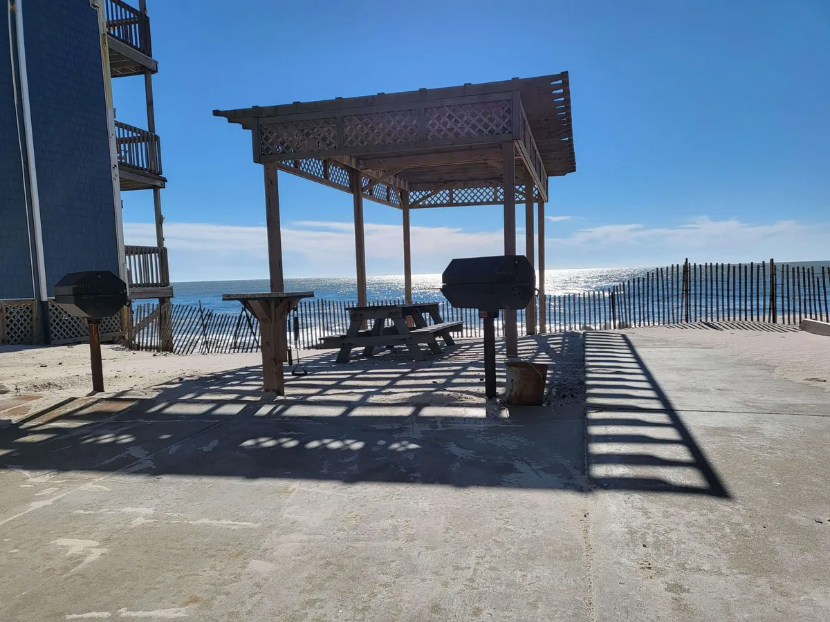 An inviting picnic table and grill set against the backdrop of an ocean view, with sandy ground and space for children to play nearby. The setup evokes images of casual family barbecues and outdoor socializing just steps from the beach.
