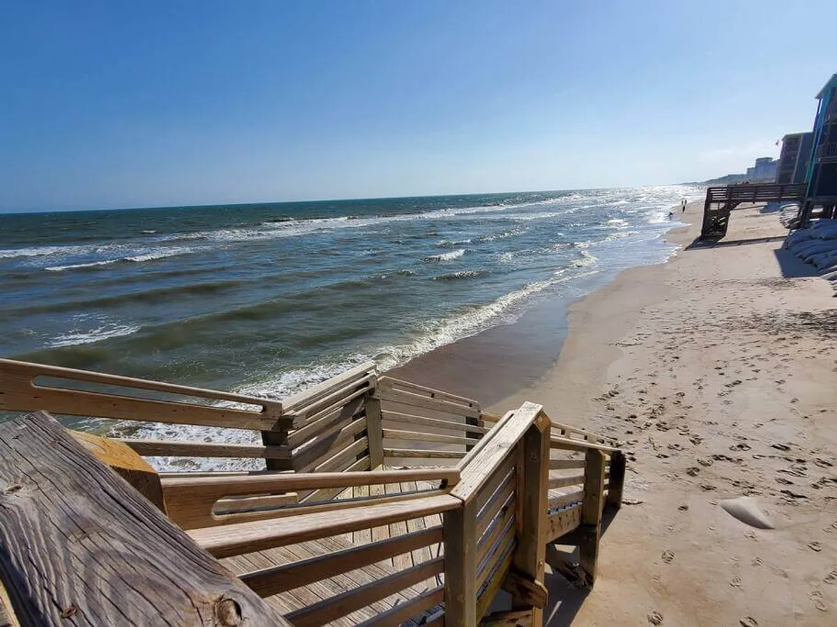 A sunlit exterior shot showcases wooden stairs leading down to a broad sandy beach, with waves rolling onto the shore and sunlight glinting across the water. The photo captures an inviting pathway to seaside fun and the potential for collecting seashells or strolling along the surf.