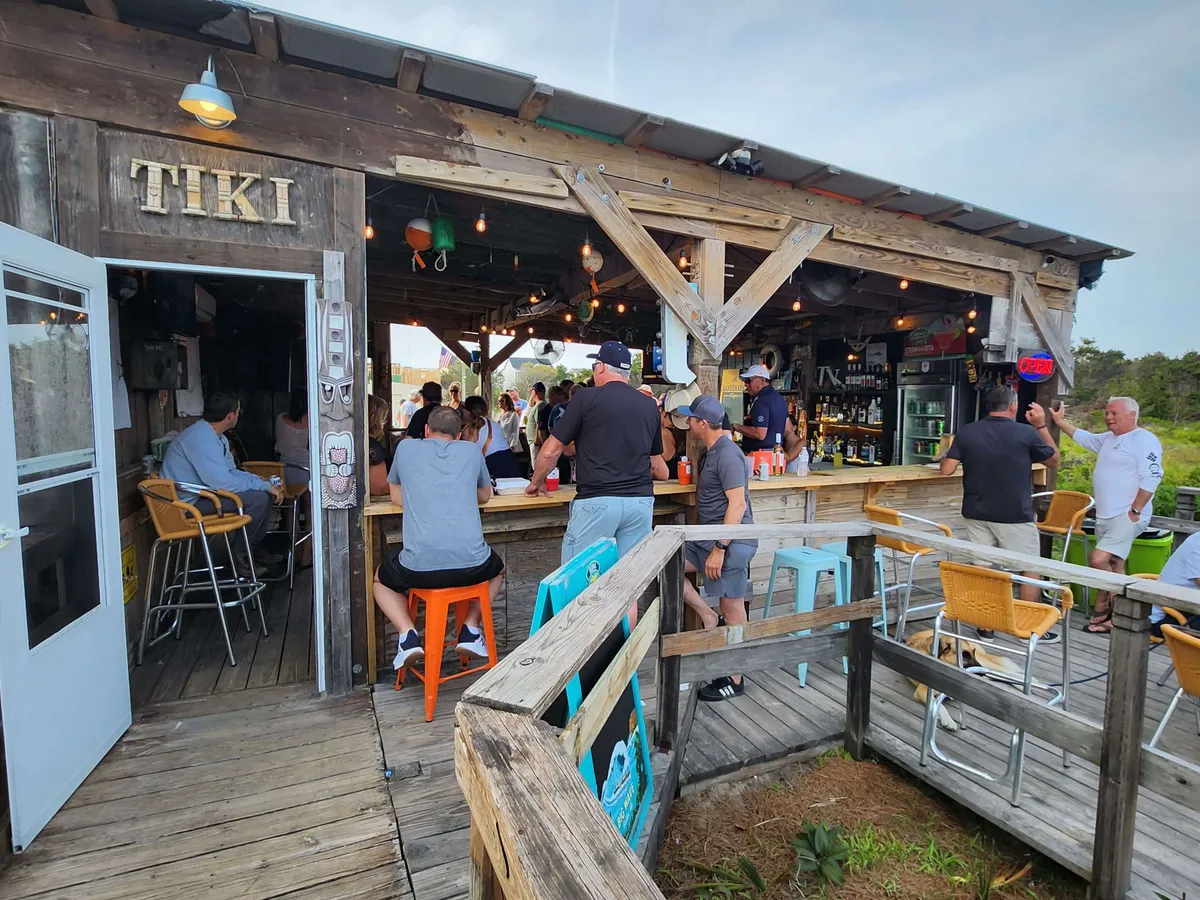 An inviting tiki bar with outdoor seating and patrons enjoying drinks, bathed in the warm glow of sunset. The festive yet relaxed atmosphere encourages socializing and unwinding after a day on the beach, echoing the idea of exploring local waterfront restaurants.