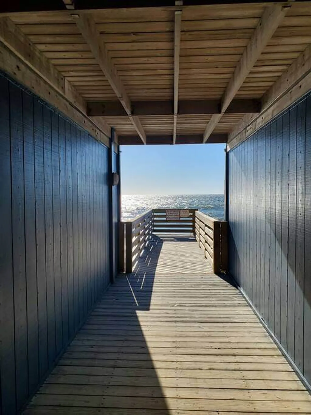 A wooden walkway leads directly towards the ocean, bordered by blue-walled buildings. The walkway invites exploration, and the ocean view beyond is calm and inviting, evoking the spirit of outdoor breaks and beachside relaxation.