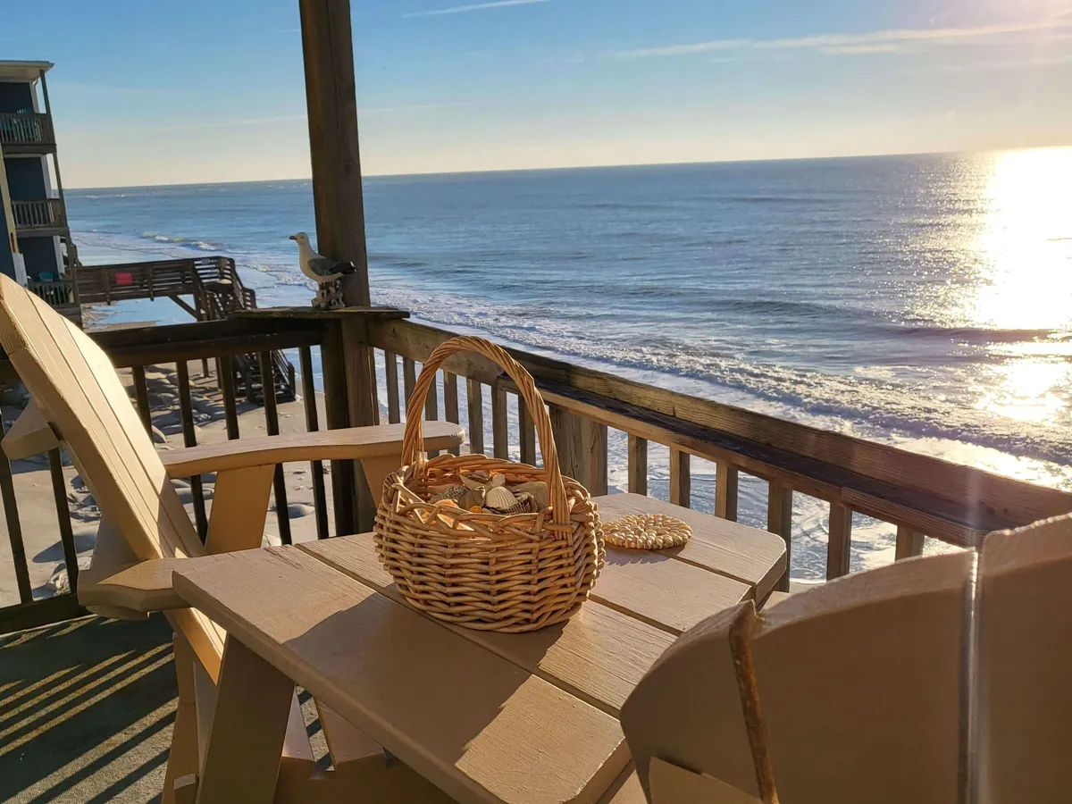 The image showcases a serene oceanfront view from the private balcony of a second-floor condo. Wooden deck chairs invite relaxation, while a wicker basket suggests leisurely mornings or sunset snacks. The tranquil sea stretches out before you, hinting at sunrise views and the soothing sound of waves.